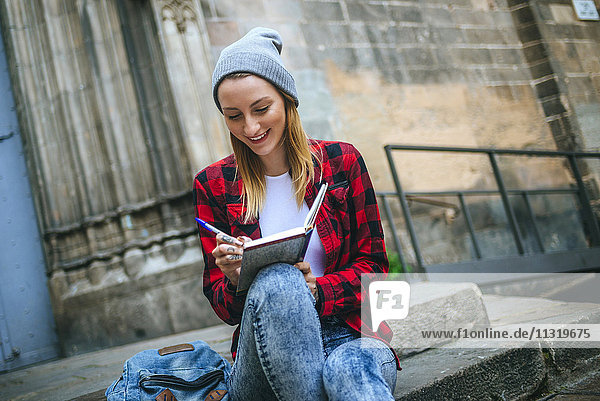 Spain  Barcelona  smiling young woman sitting on stairs writing in notebook