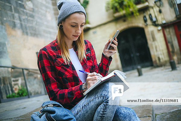 Spain  Barcelona  young woman with cell phone sitting on stairs writing in notebook