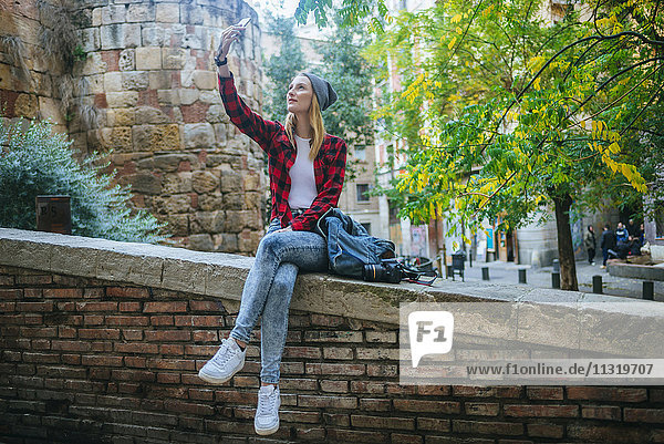 Spain  Barcelona  young woman sitting on a wall taking selfie with cell phone