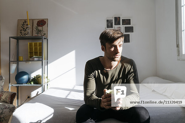 Unhappy young man sitting on bed with cup of coffee