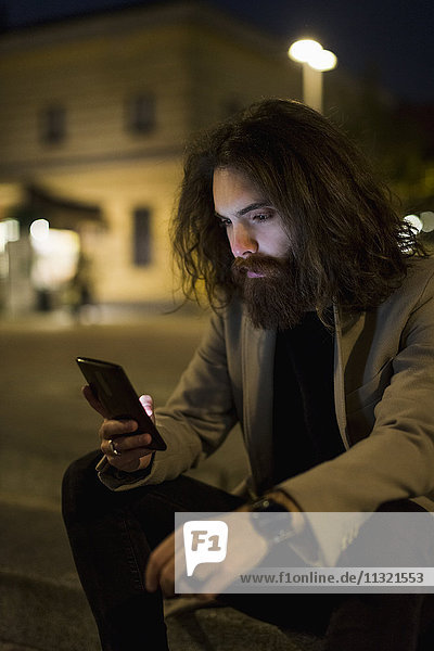 Stilvoller junger Mann im Freien in der Stadt bei Nacht mit Blick auf Handy