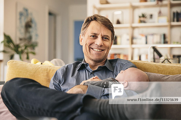Lächelnder Vater und sein neugeborenes Baby sitzen zu Hause auf der Couch.