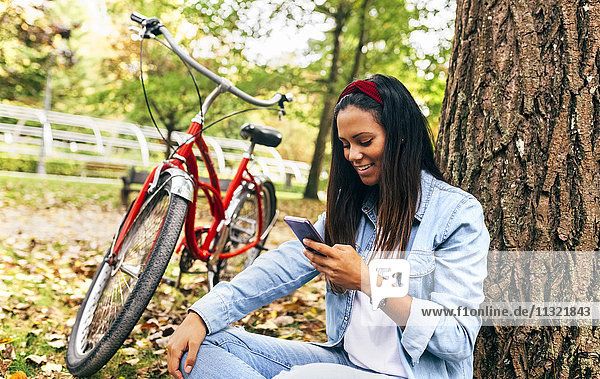 Lächelnde junge Frau schaut im Herbst auf das Handy im Park