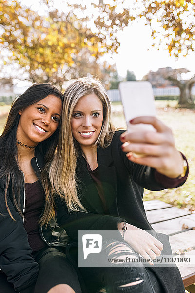 Two smiling young women on park bench taking a selfie