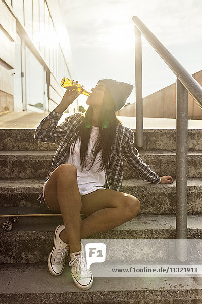 Junge Frau sitzt auf einem Skateboard und trinkt ein Bier aus der Flasche.