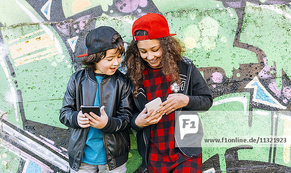 Two children standing in front of graffiti wall looking at cell phone