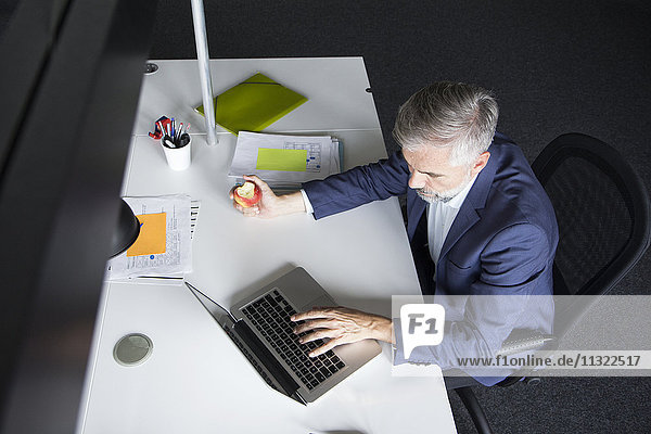 Businessman using laptop and eating an apple at office desk
