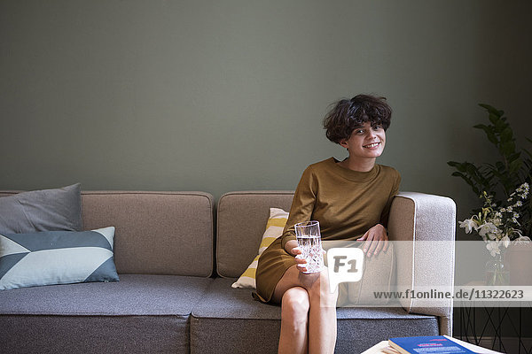 Porträt einer lächelnden jungen Frau mit einem Glas Wasser  die zu Hause auf der Couch sitzt.