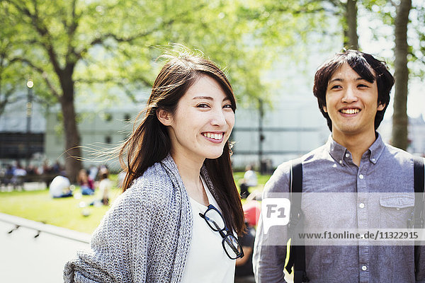 Ein junger Japaner und eine junge Japanerin genießen einen Tag in London  stehen in einem Park und lächeln.
