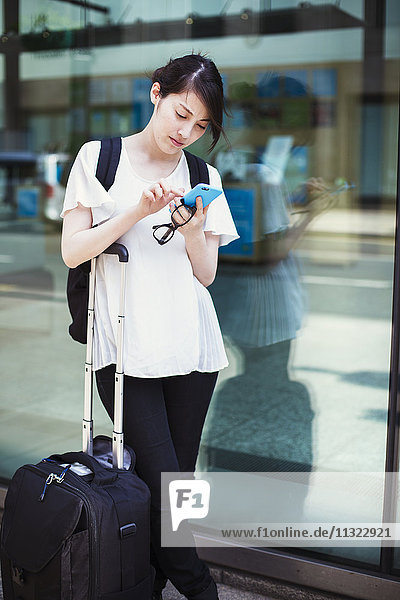 Junge Japanerin genießt einen Tag in London  steht mit einem Koffer draußen und benutzt ein Smartphone.