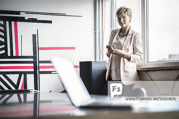 Confident businesswoman with cell phone standing in modern office