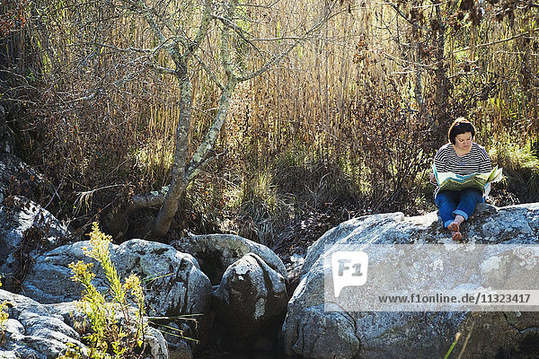 Eine Frau sitzt auf den Felsen im Schatten eines Flusses und schaut auf eine Karte.
