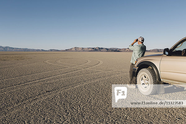 Man standing on vast desert  looking through binoculars and leaning against truck  Black Rock Desert
