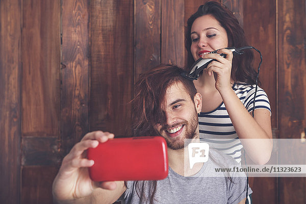 Junger Mann  der einen Selfie nimmt  während er sich von seiner Freundin mit einer Haarschneidemaschine die Haare schneiden lässt.