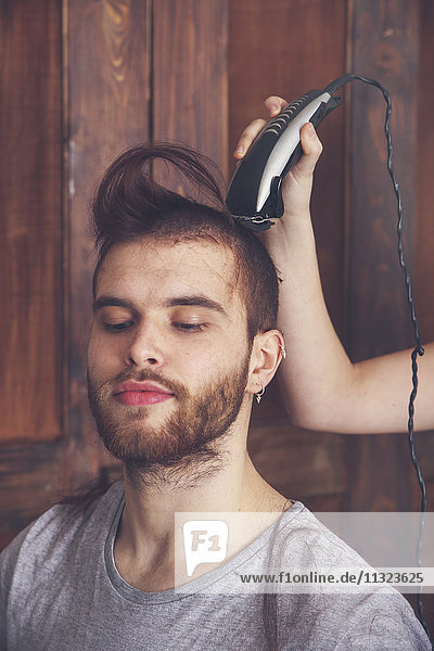 Porträt eines jungen Mannes  der sich von seiner Freundin mit einer Haarschneidemaschine frisieren lässt