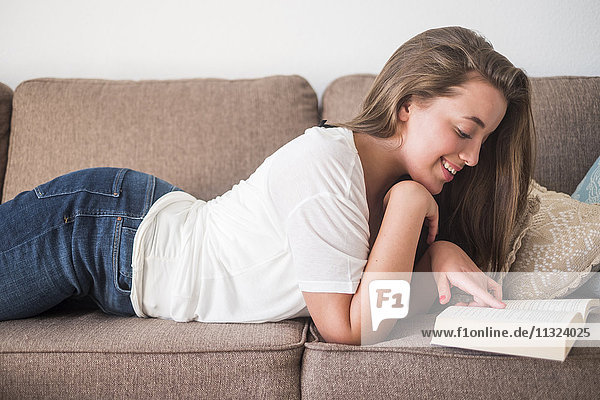 Lächelnde junge Frau auf der Couch liegend Lesebuch
