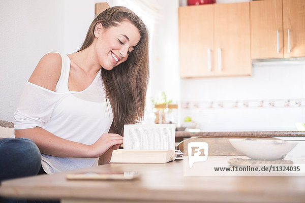 Lächelnde junge Frau sitzt am Tisch in der Küche und liest Buch.