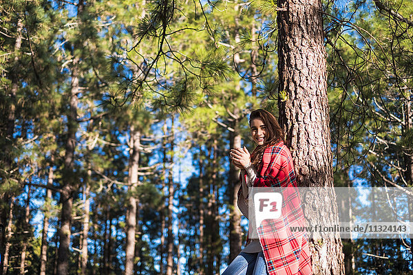 Lächelnde junge Frau mit Decke und Getränk an Baumstamm gelehnt im Wald
