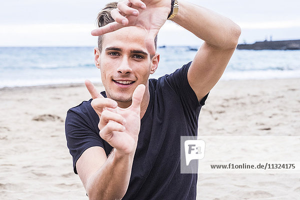 Porträt eines lächelnden jungen Mannes am Strand  der mit den Fingern den Rahmen baut