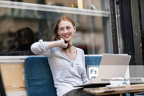 Lächelnde rothaarige Frau mit Laptop im Straßencafé