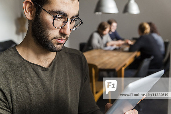 Junger Mann mit digitalem Tablett im modernen Büro mit Mitarbeitern am Tisch im Hintergrund