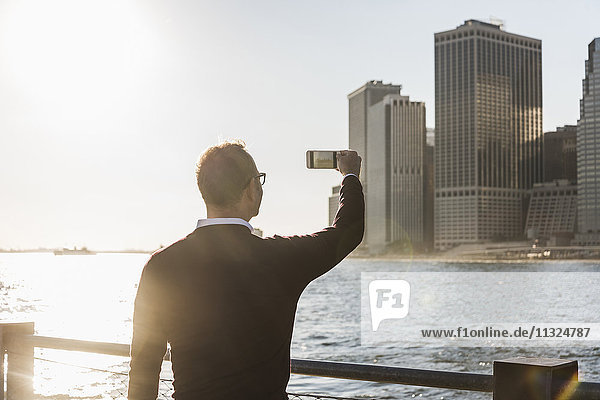 USA  Brooklyn  Mann fotografiert Manhattan Wolkenkratzer mit Smartphone in der Abenddämmerung