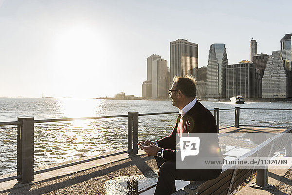 USA  Brooklyn  entspannter Mann mit Smartphone auf der Bank sitzend  mit Blick auf die Aussicht