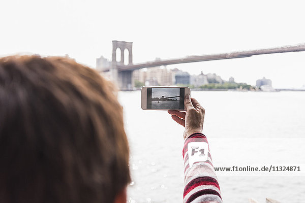 USA  New York City  Frau am East River macht Handyfoto von der Brooklyn Bridge