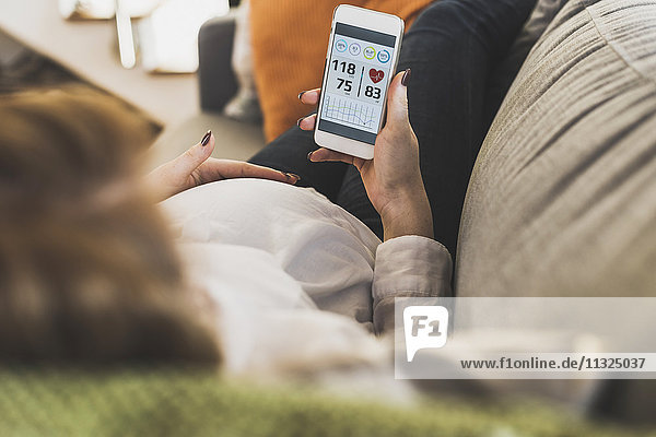 Schwangere Frau auf der Couch mit Smartphone zur Überprüfung der Gesundheitsdaten