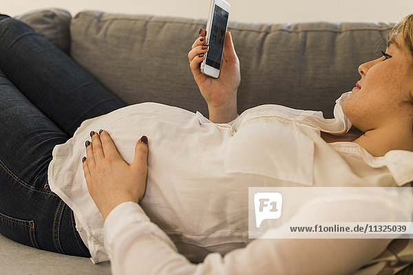 Schwangere Frau auf der Couch mit Smartphone zur Überprüfung der Gesundheitsdaten