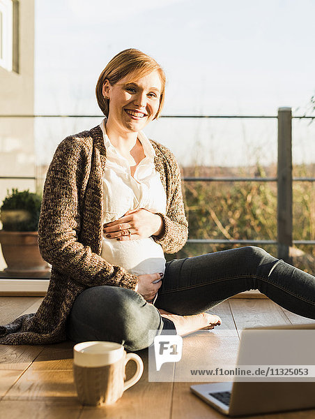 Lächelnde schwangere Frau auf dem Boden sitzend mit Laptop und Becher