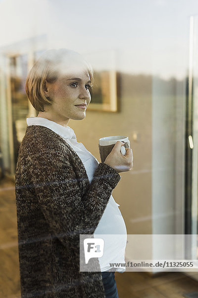 Schwangere Frau hinter der Fensterscheibe mit einem Drink im Becher