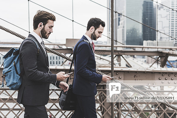 USA  New York City  two businessmen with cell phones and earbuds on Brooklyn Bridge