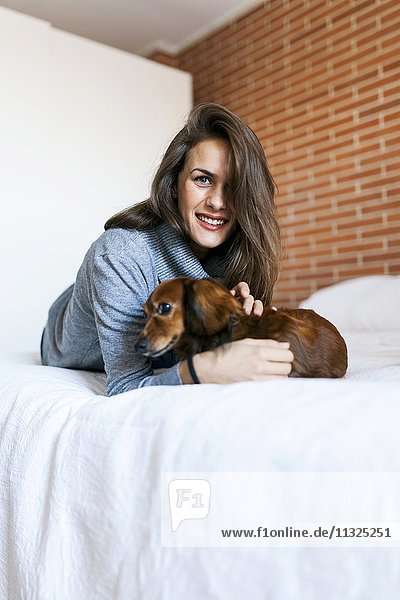 Lächelnde junge Frau mit ihrem Hund im Bett liegend