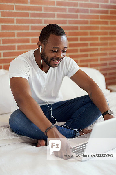 Smiling young man with earphones sitting on bed using laptop