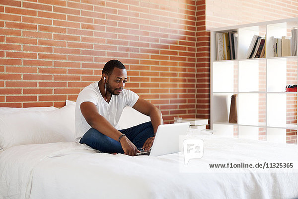 Junger Mann mit Kopfhörern auf dem Bett sitzend mit Laptop