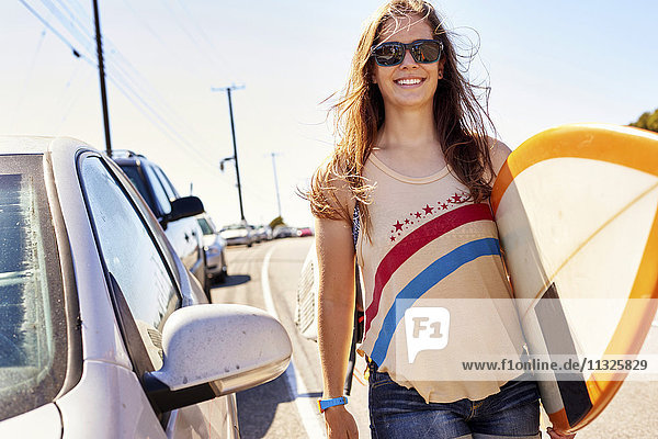 Lächelnde junge Frau mit Surfbrett auf der Küstenstraße