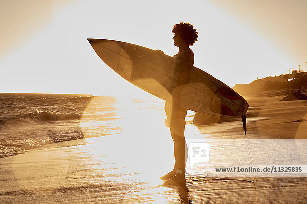 Junger Mann mit Surfbrett am Strand bei Sonnenuntergang