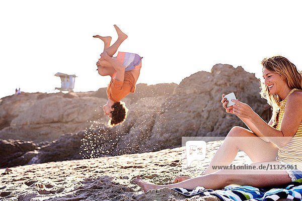 Junger Mann am Strand beim Salto mit Frau beim Handy-Checken