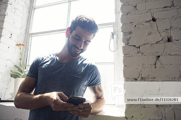 Lächelnder junger Mann mit Blick auf das Handy am Fenster