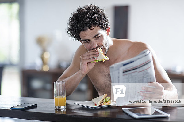 Young man at home reading newspaper and eating sandwich