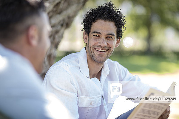 Smiling young man holding book looking at friend in park