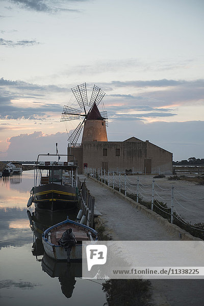 Boats moored in canal by traditional windmill against sky during sunset