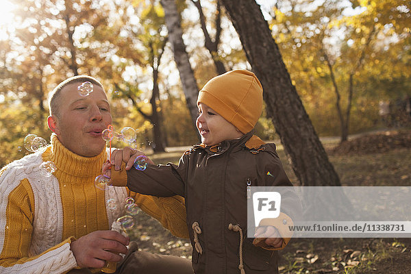 Father blowing bubbles with baby boy in park during autumn