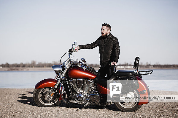 Full length portrait of biker standing by motorcycle and looking away at lakeshore