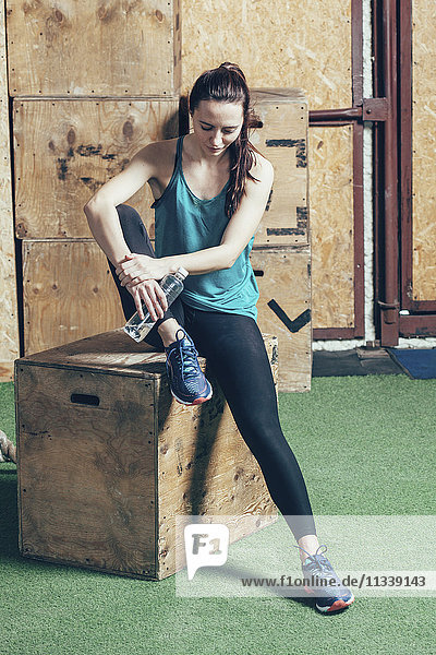 Full length of female athlete holding water bottle while sitting on wooden box at gym