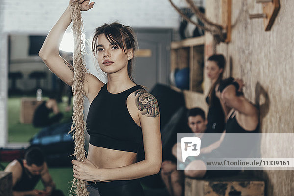 Portrait of confident female athlete holding rope while standing at gym with friends in background