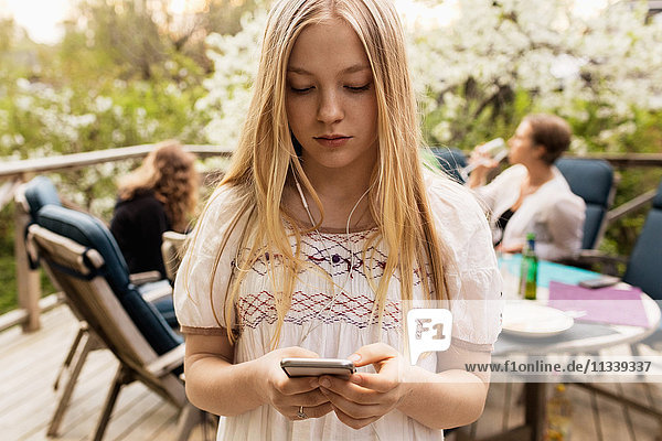 Teenager-Mädchen mit Smartphone im Hof mit Familie im Hintergrund