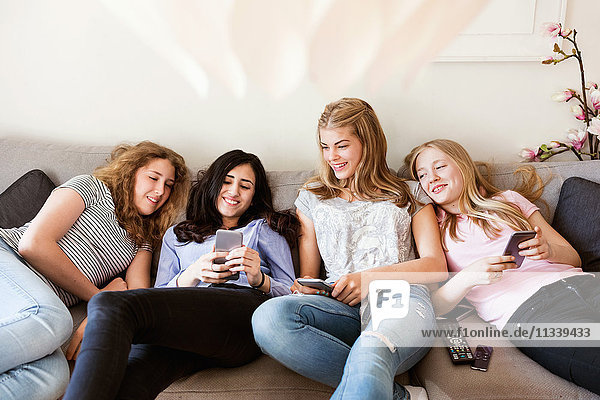 Happy teenage girls using smart phone while reclining on sofa in living room