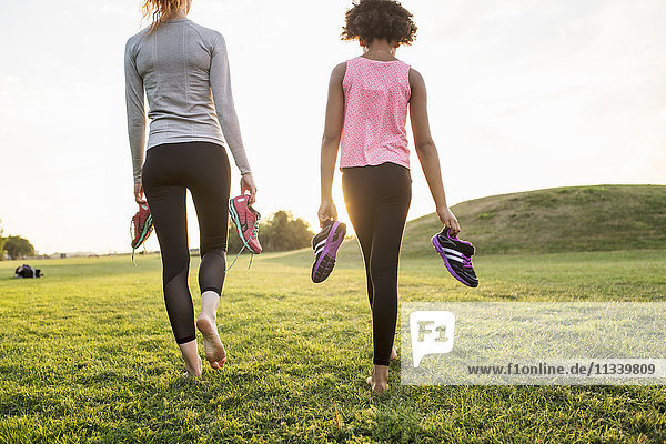 Rear view of mother and daughter holding sports shoes while walking on grass at park during sunset